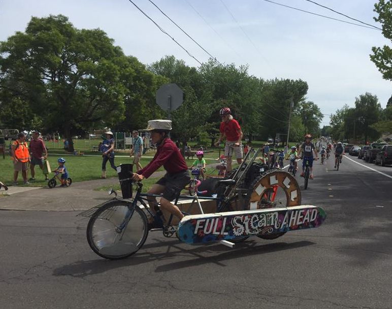 A person rides a kinetic sculpture bike that says "Full Steam Ahead"