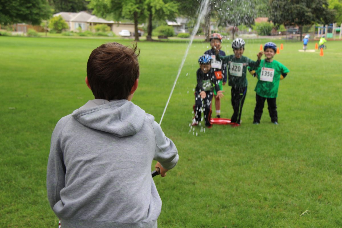Kids wearing bicycle helmets get sprayed by water.