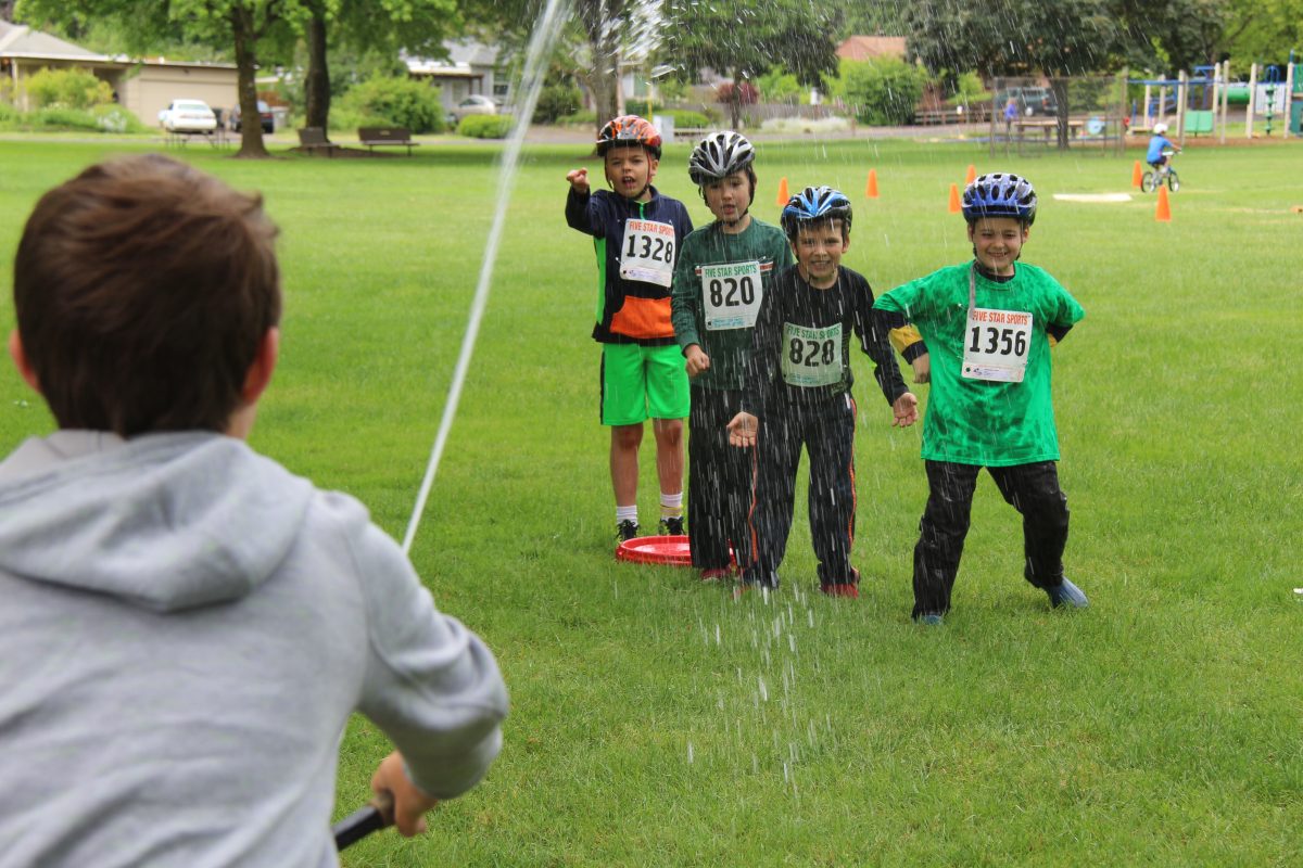 Kids wearing bicycle helmets get sprayed by water.