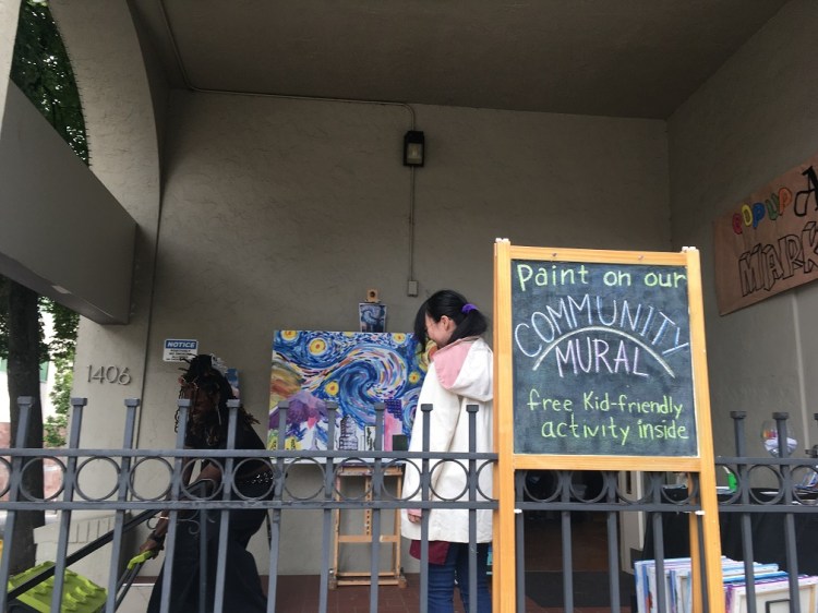 Sign says "Paint on our community mural" with large, painted canvas in background